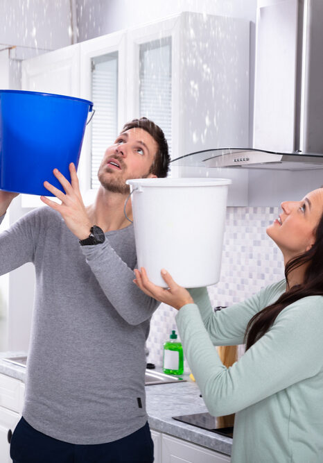Close-up Of A Worried Young Couple Collecting Water Leaking From Ceiling In The Blue Bucket